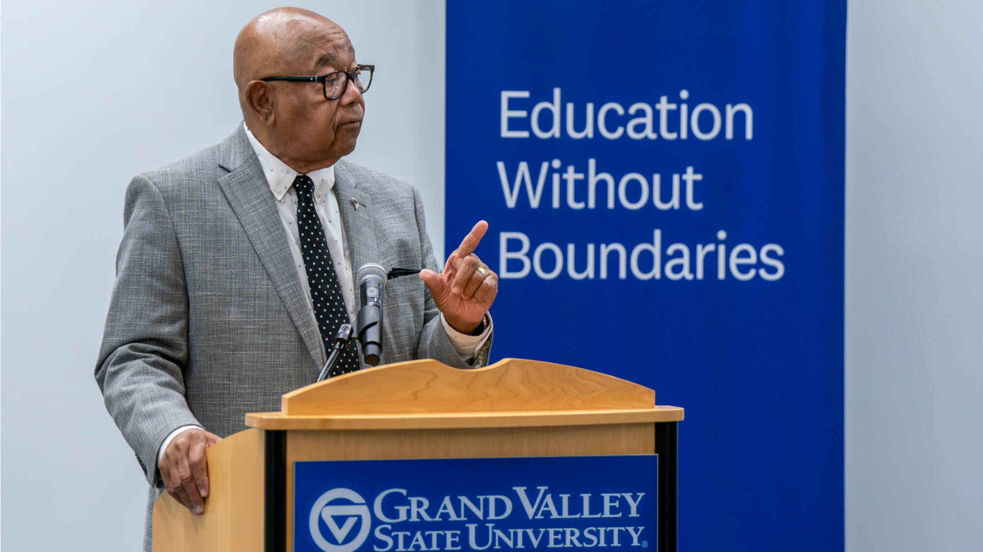 A man speaks at a podium with a Grand Valley State University logo and a blue banner in the background.
